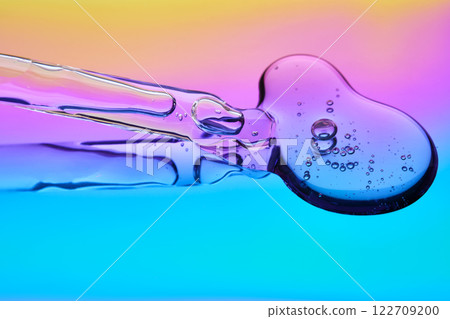 A drop of cosmetic serum and a pipette on a multicolored background in shades of blue and lilac. A drop of cosmetic serum and a pipette on a multicolored background in shades of blue and lilac. 122709200