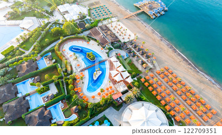 Aerial view of a beachfront luxury resort with a wave-shaped pool, private pier, and organized sunbathing areas 122709351