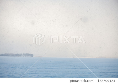Snow is gently falling on a vast frozen lake on a cold winter day, creating a serene and peaceful atmosphere with a distant island covered in trees 122709423