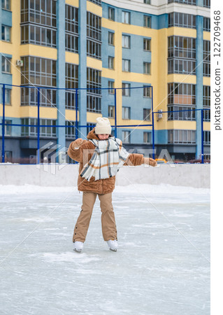 Girl wearing warm winter clothes ice skating on a rink in front of a residential building, enjoying the thrill of winter sports and embracing a fun outdoor leisure activity 122709468