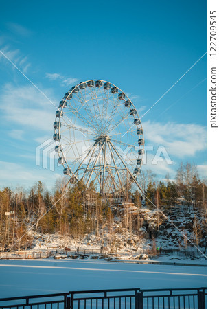 Ferris wheel standing tall against a clear blue winter sky, with snow-covered trees and rocky terrain in the foreground, creating a picturesque winter scene Ferris wheel standing tall against a clear blue winter sky, with snow-covered trees and rocky terrain in the foreground, creating a picturesque winter scene 122709545