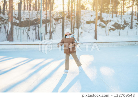 Happy girl wearing winter clothes ice skating outdoors on a sunny winter day in a snowy forest, enjoying healthy lifestyle and having fun during winter holidays Happy girl wearing winter clothes ice skating outdoors on a sunny winter day in a snowy forest, enjoying healthy lifestyle and having fun during winter holidays 122709548