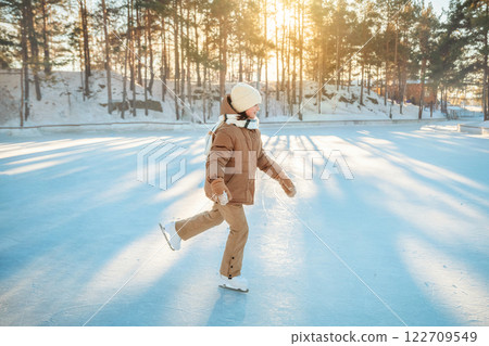Happy girl wearing winter clothes ice skating outdoors on a sunny winter day in a snowy forest, enjoying healthy lifestyle and having fun during winter holidays Happy girl wearing winter clothes ice skating outdoors on a sunny winter day in a snowy forest, enjoying healthy lifestyle and having fun during winter holidays 122709549