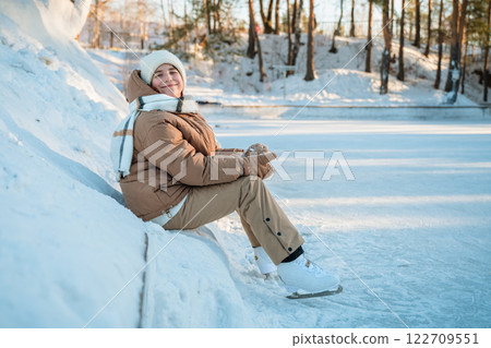 cute girl wearing winter clothes sitting on snowbank by frozen lake, resting after ice skating in beautiful winter forest, enjoying winter sports and outdoor activities on sunny day 122709551