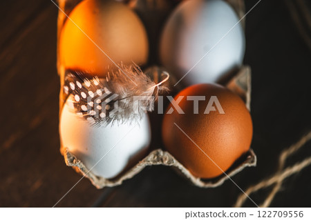 Brown and white chicken eggs are nestled in a cardboard carton, a speckled feather resting gently atop one, creating a rustic, farm-fresh still life Brown and white chicken eggs are nestled in a cardboard carton, a speckled feather resting gently atop one, creating a rustic, farm-fresh still life 122709556