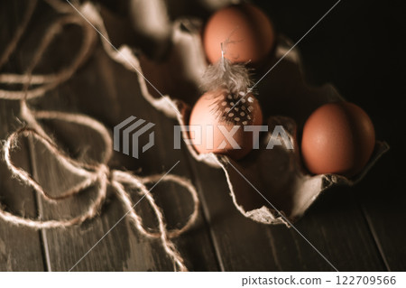 Fresh brown eggs sit in a cardboard carton on a rustic wooden table, one adorned with a delicate guinea fowl feather, creating a simple yet elegant composition, top view Fresh brown eggs sit in a cardboard carton on a rustic wooden table, one adorned with a delicate guinea fowl feather, creating a simple yet elegant composition, top view 122709566