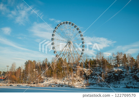 Ferris wheel standing tall against a clear blue winter sky, with snow-covered trees and rocky terrain in the foreground, creating a picturesque winter scene Ferris wheel standing tall against a clear blue winter sky, with snow-covered trees and rocky terrain in the foreground, creating a picturesque winter scene 122709570