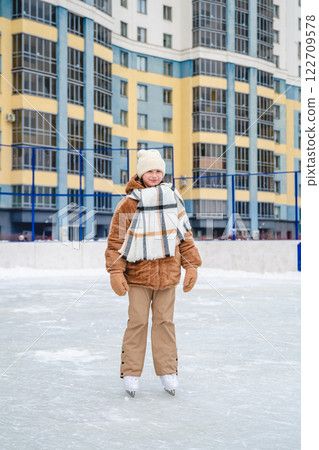 Girl wearing warm winter clothes ice skating on a rink in front of a residential building, enjoying the thrill of winter sports and embracing a fun outdoor leisure activity Girl wearing warm winter clothes ice skating on a rink in front of a residential building, enjoying the thrill of winter sports and embracing a fun outdoor leisure activity 122709578