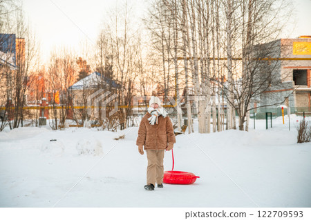 Warmly dressed girl pulling an inflatable snow tube while walking along a snowy path in a winter park, eagerly preparing for a day filled with fun sledding and winter activities Warmly dressed girl pulling an inflatable snow tube while walking along a snowy path in a winter park, eagerly preparing for a day filled with fun sledding and winter activities 122709593