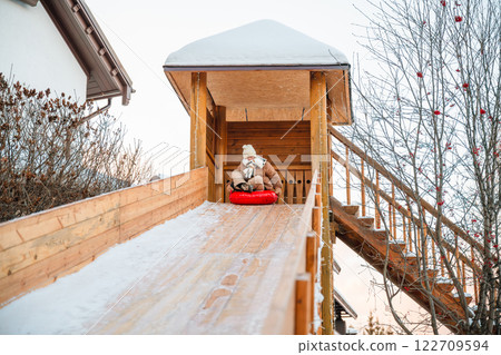 Child is enjoying a snowy day, sliding down a wooden slide on a red snow tube at a winter park, surrounded by snow-covered trees and benches 122709594