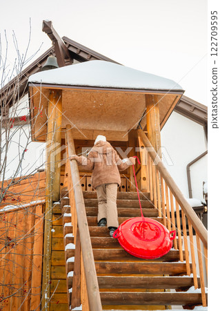 Winter scene showing a girl climbing snowy wooden stairs, pulling a red snow tube behind her, at a ski resort, ready for some fun in the snow 122709595