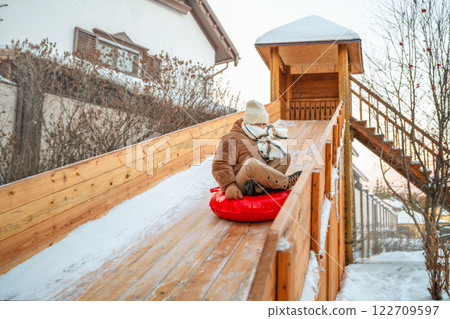 Child is enjoying a snowy day, sliding down a wooden slide on a red snow tube at a winter park, surrounded by snow-covered trees and benches Child is enjoying a snowy day, sliding down a wooden slide on a red snow tube at a winter park, surrounded by snow-covered trees and benches 122709597