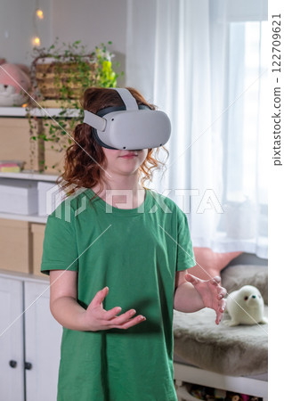redhead teen girl at home in living room put on vr helmet to enjoy virtual reality online game cyber space world simulation. cute girl has fun using new electronic device for entertainment redhead teen girl at home in living room put on vr helmet to enjoy virtual reality online game cyber space world simulation. cute girl has fun using new electronic device for entertainment 122709621