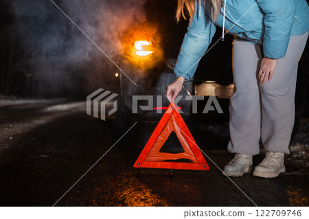 On a dark winter road, a woman places a reflective warning triangle beside her broken-down car. The scene highlights the importance of safety precautions during nighttime travel. On a dark winter road, a woman places a reflective warning triangle beside her broken-down car. The scene highlights the importance of safety precautions during nighttime travel. 122709746