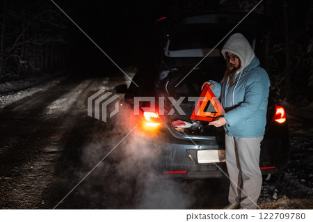 On a snowy night road, a woman sets a warning triangle after her car breaks down in a winter forest. Her car's trunk is open, and steam rises from the cold, creating a dramatic scene. On a snowy night road, a woman sets a warning triangle after her car breaks down in a winter forest. Her car's trunk is open, and steam rises from the cold, creating a dramatic scene. 122709780