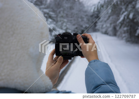 A photographer holds a digital camera and takes pictures of snow-covered trees and a winding path in a serene winter forest landscape. A woman takes a photo as a keepsake of a winter forest. A photographer holds a digital camera and takes pictures of snow-covered trees and a winding path in a serene winter forest landscape. A woman takes a photo as a keepsake of a winter forest. 122709787