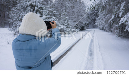 A woman takes a photo of a fabulous winter forest landscape. A photographer takes pictures of a snowy road winding through a winter forest, surrounded by falling snowflakes and peaceful landscapes. 122709791