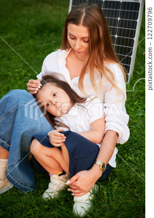 Mother and daughter sit together on grass, embracing in warm moment with solar panel beside in the evening. Concept of renewable energy and family, highlighting commitment to sustainable future. 122709796