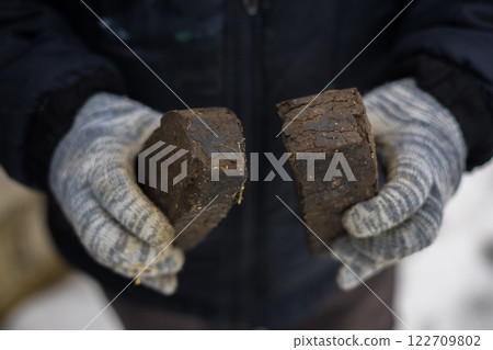A large plan of peat briquettes for kindling a stove in hands. A farmer in gloves holds two peat briquettes for kindling a stove in winter, which is a sustainable and traditional heating solution. 122709802