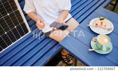 Cropped photo of girl putting phone on charge from solar battery. Young girl relaxing in cafe while charging smartphone from solar battery charger. Concept of alternative energy and green lifestyle. Cropped photo of girl putting phone on charge from solar battery. Young girl relaxing in cafe while charging smartphone from solar battery charger. Concept of alternative energy and green lifestyle. 122709807