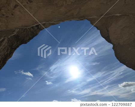 Bright sun shining through rocky arch with blue sky and wispy clouds situated in a natural landscape 122709887