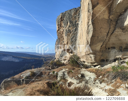 Expansive view of ancient caves carved into cliffs in a dramatic mountainous landscape under a clear blue sky Expansive view of ancient caves carved into cliffs in a dramatic mountainous landscape under a clear blue sky 122709888