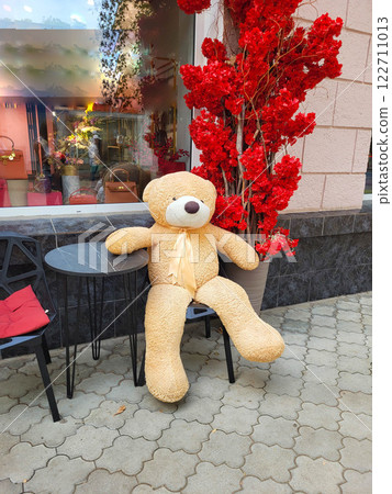 A large light-colored teddy bear sits on a chair in an outdoor cafe A large light-colored teddy bear sits on a chair in an outdoor cafe 122711013