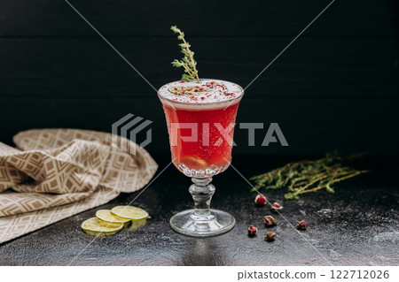 Elegant red cocktail with lime, herbs, and floral garnish, served in vintage glass on dark surface 122712026