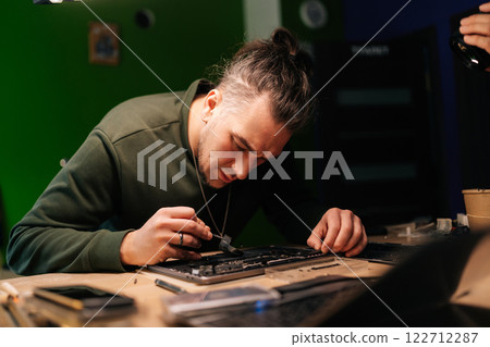 Portrait of electronic technician inspecting damaged laptop motherboard, diagnosing complex hardware malfunction sitting at table in professional repair workspace. Process electronic device repair. 122712287