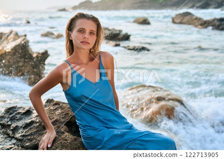 Model woman sits on cliff in wet dress at exotic island ocean. Stunning femininity, divine beauty with storming waves. Perfect for travel, nature, fashion themes. Female model, elegance by sea. Model woman sits on cliff in wet dress at exotic island ocean. Stunning femininity, divine beauty with storming waves. Perfect for travel, nature, fashion themes. Female model, elegance by sea. 122713905