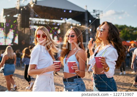 Group of happy young women with sunglasses enjoy summer music festival on sandy beach. Holding colorful drinks, dancing with stage in background. Casual outfits, trendy accessories, lively crowd. Group of happy young women with sunglasses enjoy summer music festival on sandy beach. Holding colorful drinks, dancing with stage in background. Casual outfits, trendy accessories, lively crowd. 122713907