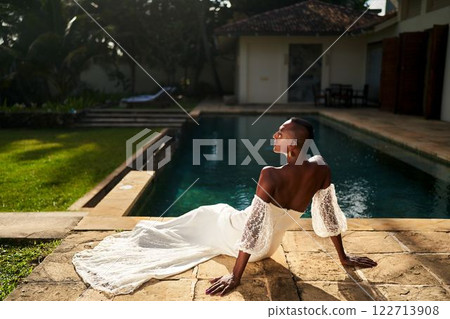 Person in white wedding dress by pool. Gender fluid individual enjoys sunlit moment. LGBTQ wedding, modern bridal fashion, exotic vacation. Tranquil luxury villa, elegant identity celebration. 122713908