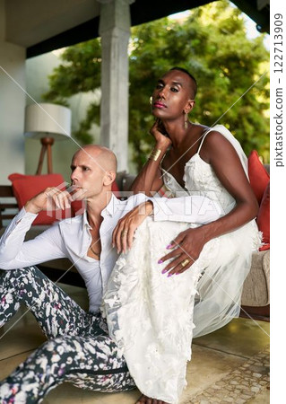 Gender fluid person in white bridal dress, jewelry, sits beside LGBTQ groom in patterned suit. Wedding at tropical villa. Diverse marriage. Inclusive fashion for modern couples. 122713909