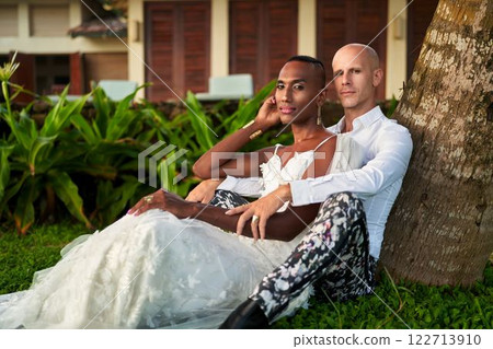 Bride in white dress and groom in bold suit at tropical villa. LGBTQ wedding theme with a gender fluid black person and groom in vibrant outfit. Celebrating love and diversity in exotic setting. 122713910
