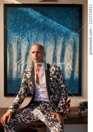 Bald man in bold floral suit sits at villa, possible groom outfit. Elegant gay fashion, unique style for weddings. Trendy chic look, modern expression. Luxury setting indoors, artistic blue painting. 122713911