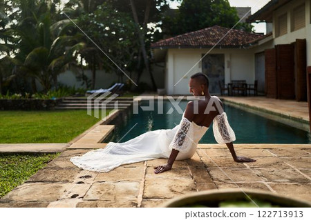 Black person in wedding dress at villa. Silk gown by poolside, green garden. Celebrating pride, embracing identity, LGBTQ couple love. Trendy bridal elegance in exotic modern venue. Inclusive wedding. Black person in wedding dress at villa. Silk gown by poolside, green garden. Celebrating pride, embracing identity, LGBTQ couple love. Trendy bridal elegance in exotic modern venue. Inclusive wedding. 122713913