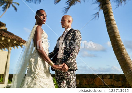 Gender fluid couple holds hands in wedding at tropical villa. Bride in white dress, groom in floral suit. LGBTQ theme. Ceremony at sunny paradise location. 122713915