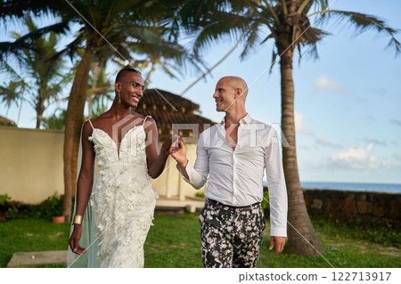 Ethnic black person in white dress with LGBTQ groom in bold suit at tropical villa wedding. Ambiguous gender fluid bride, groom in beautiful ceremony. Exotic palm tree setting. 122713917