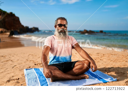 Senior man with stylish beard and sunglasses meditates on beach. Older male sits cross-legged on towel by ocean waves. Wellness, relaxation, mindfulness for active elderly. 122713922