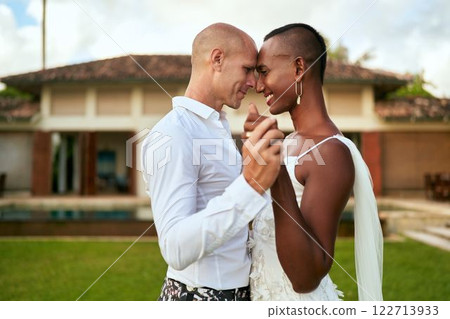 LGBTQ couple dances in villa wedding. Bride in white dress, groom in bold suit. Tropical background creates romantic atmosphere. Joyful celebration scene, inclusive love. 122713933