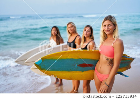 Young women with surfboards on beach. Friends enjoy summer surfing together. Female surfers in bikinis, vibrant energy, celebrate International Women Day through sport, adventure. Young women with surfboards on beach. Friends enjoy summer surfing together. Female surfers in bikinis, vibrant energy, celebrate International Women Day through sport, adventure. 122713934