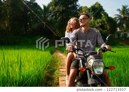 Couple rides motorbike on rural path by rich rice fields. Man drives scooter with woman enjoying green scenery. Romantic adventure amid nature. Explore countryside, feel freedom. Travel concept. 122713937
