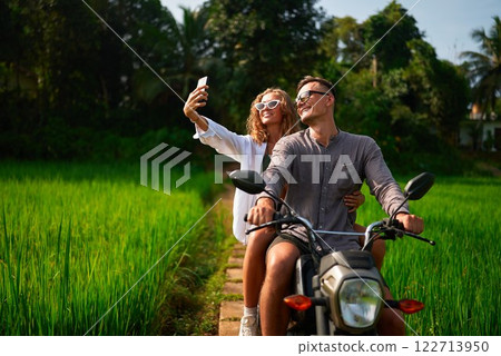 Happy couple on motorbike taking selfie in lush green rice fields. Smiling girl with curly hair, white sunglasses holds phone. Man in casual clothes drives scooter through scenic tropical landscape. Happy couple on motorbike taking selfie in lush green rice fields. Smiling girl with curly hair, white sunglasses holds phone. Man in casual clothes drives scooter through scenic tropical landscape. 122713950