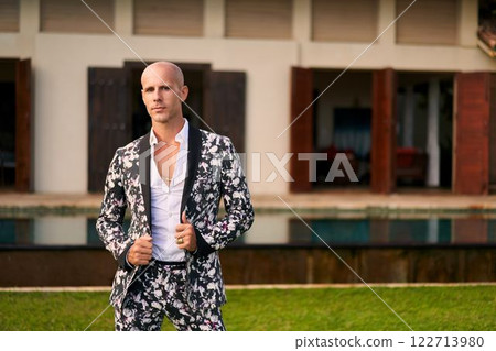 Bald man, bold floral suit poses villa courtyard. Stylish outfit, modern groom trends. Luxury poolside. Elegant attire, fashion statement. Perfect for weddings, upscale events, fashion, LGBTQ. 122713980