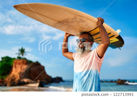 Senior black man with surfboard on sunny ocean beach holds board overhead. Active older male surfs waves in sea, enjoys coastal lifestyle. Fit elder engages in sporty adventure, healthy retirement. Senior black man with surfboard on sunny ocean beach holds board overhead. Active older male surfs waves in sea, enjoys coastal lifestyle. Fit elder engages in sporty adventure, healthy retirement. 122713991