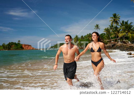 Fit couple runs on tropical beach, hand in hand, splashing water. Athletic man and slender woman in swimwear enjoy active vacation, jogging by sea. Exercising together, they embody wellness, travel. 122714012