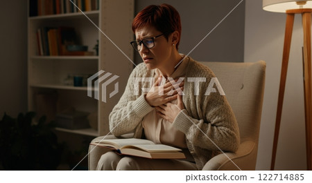 Mature woman experiencing discomfort at home, holding chest, surrounded by books and cozy atmosphere Mature woman experiencing discomfort at home, holding chest, surrounded by books and cozy atmosphere 122714885
