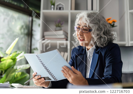 Elderly female business executive analyzing reports and making notes in modern office with green plants 122716703