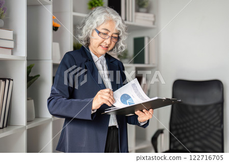 Elderly business woman analyzing reports and checking documents on a clipboard in an organized office) 122716705