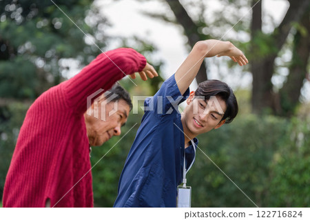 Young caregiver encouraging elderly man to perform arm stretching exercise in garden Young caregiver encouraging elderly man to perform arm stretching exercise in garden 122716824